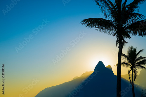 Scenic sunset view of Two Brothers Mountain backlit by the setting sun with palm tree silhouettes lining Ipanema Beach in Rio de Janeiro  Brazil