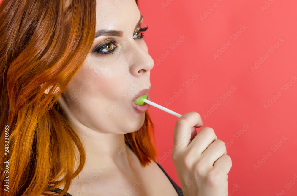 Portrait of a cheerful beautiful woman with red hair licking caramel green candy on a stick over a red background. Joy, sweets, candy. Entertainment concept Portrait of a cheerful beautiful woman with red hair licking caramel green candy on a stick over a red background. Joy, sweets, candy. Entertainment concept