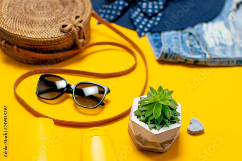 Female summer fashion background. Clothes and accessories on yellow background. Blue hat, denim shorts, round rattan bag, sandals, sunglasses, sunscreen. Flat lay top view beauty fashion blog concept