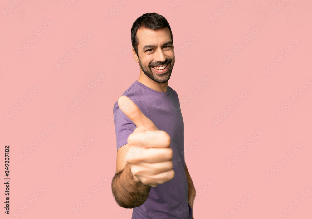 Handsome man giving a thumbs up gesture because something good has happened on isolated pink background