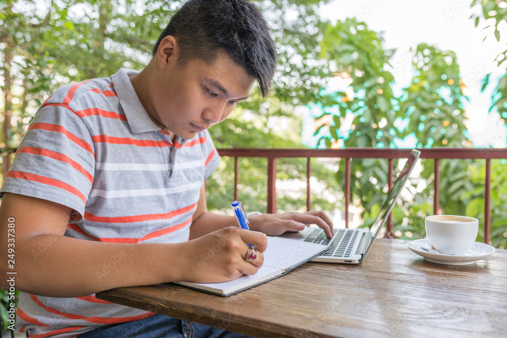 Asian man writing on notebook or document and using laptop Asian man writing on notebook or document and using laptop