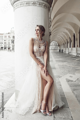 Beautiful bride in elegant, luxury ivory dress and messy updo hairstyle, posing near column in Venice, Italy
