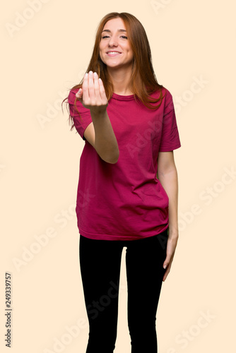 Young redhead girl inviting to come with hand. Happy that you came on isolated yellow background