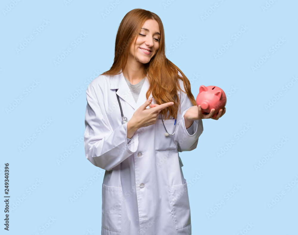 Redhead doctor woman holding a piggybank on isolated blue background Redhead doctor woman holding a piggybank on isolated blue background