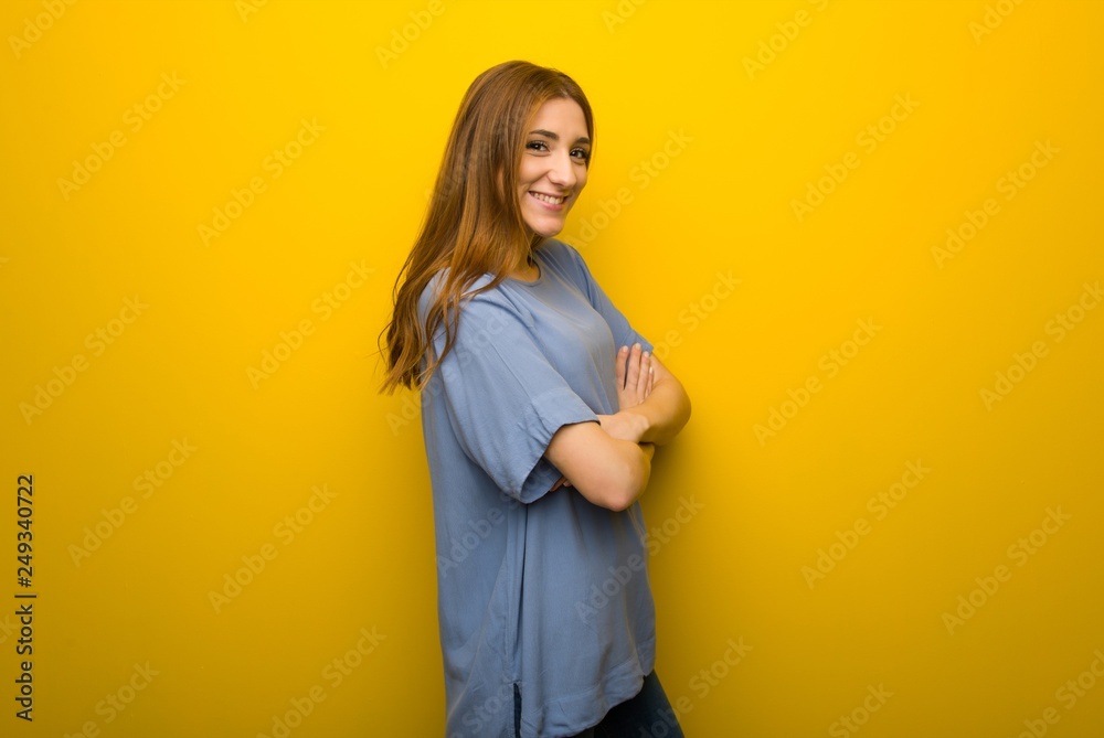 Obraz premium Young redhead girl over yellow wall background keeping the arms crossed in lateral position while smiling