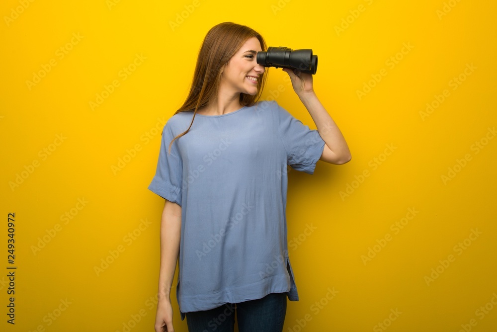 Young redhead girl over yellow wall background and looking in the distance with binoculars Young redhead girl over yellow wall background and looking in the distance with binoculars