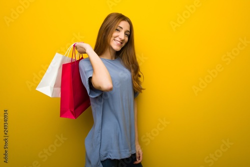 Young redhead girl over yellow wall background holding a lot of shopping bags