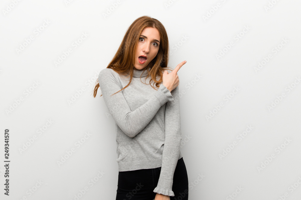 Redhead girl over white wall surprised and pointing side Redhead girl over white wall surprised and pointing side