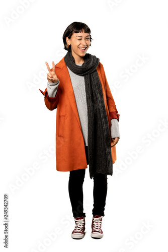 A full-length shot of a Short hair woman with coat smiling and showing victory sign over isolated white background