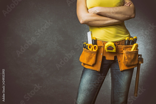 Handywoman with folded arms and tool belt on concrete background.