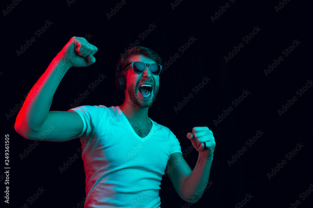 Enjoying his favorite music. Happy young stylish man in sunglasses with headphones listening sound and smiling while standing against blue neon background Enjoying his favorite music. Happy young stylish man in sunglasses with headphones listening sound and smiling while standing against blue neon background