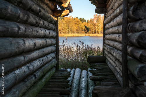 View to the tranquil blue lake, orange autumn trees and reeds through the narrow lane between two old log huts of ancient Lake Castle in Araisi Archaeological Museum Park, Latvia. photo