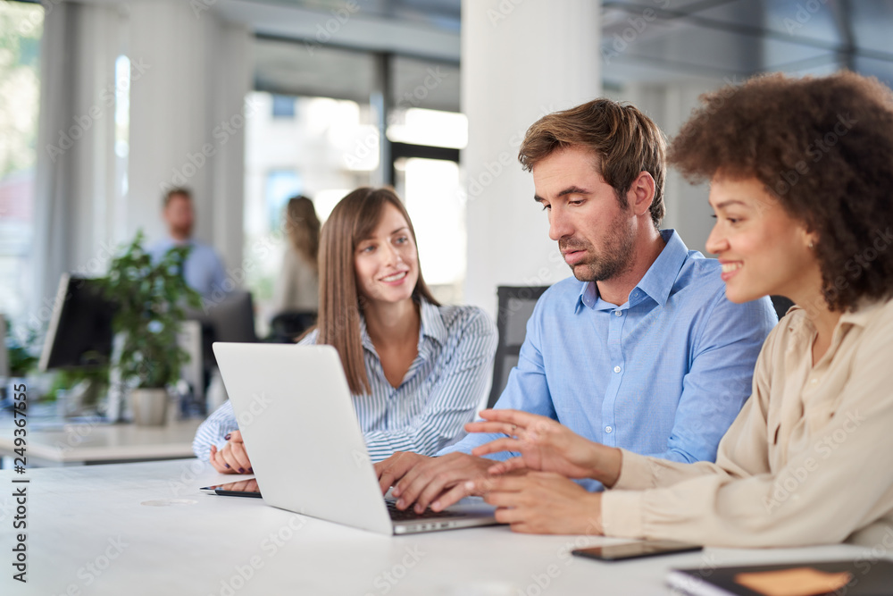 Coworkers sitting at table and solving problem. Man using laptop while two female colleagues looking at laptop and helping him.. Coworkers sitting at table and solving problem. Man using laptop while two female colleagues looking at laptop and helping him..