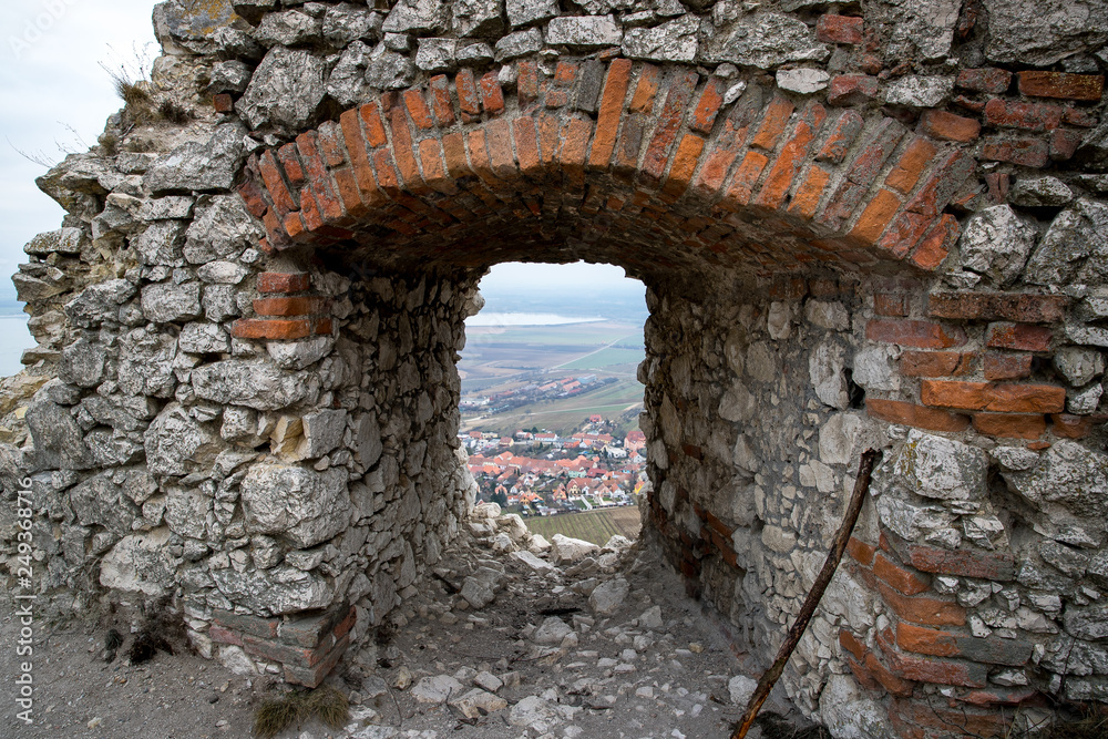 A view across the large brick window of an old castle in a village that lies in the background of a large hill
