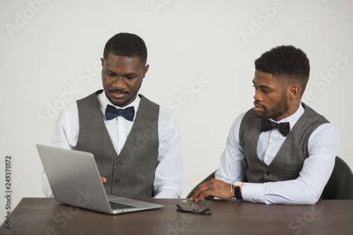 two stylish african men in suits are sitting at the table with a laptop on a white background