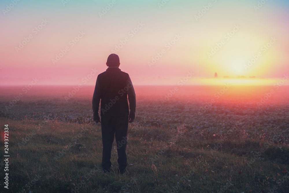 A man stands in the field in the early morning and looks at the sunrise