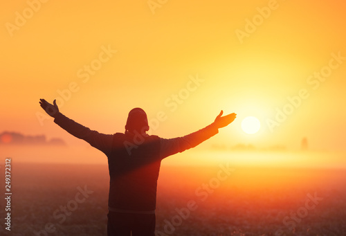 A man with his hands in the air stands in the field in the early morning and looks at the sunrise