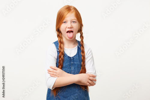 Daughter does not want to obey her parents, grimaces at the camera shows tongue, stands with her hands crossed, shows insult, dressed in denim overalls,red hair braided, isolated on white background