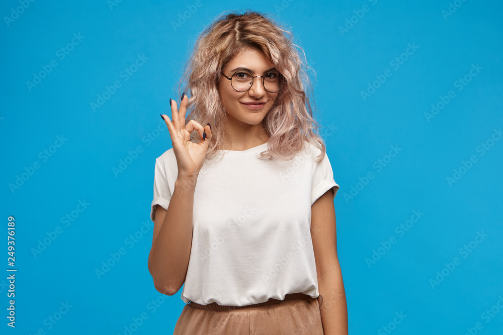 Fototapeta premium Positive friendly looking young woman with curly pinkish hair looking at camera with cute confident smile, making ok gesture. Pretty girl in round eyewear saying that everything is under control