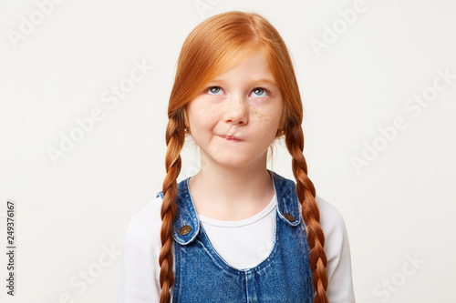 Serious little girl with freckles and braided in two long plaits red hair thinks, looks up incredulously, ponders about kindergarten, with doubt, one corner of the lips pursed, white background photo