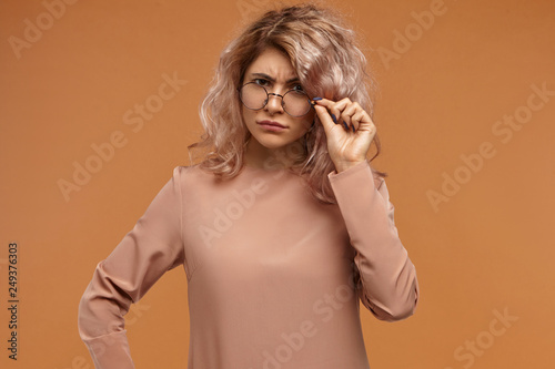 What did you mean? Studio shot of strict fashionable young European female teacher with curly hair frowning and lowering her stylish eyewear, staring at camera with disapproval or dispraise photo