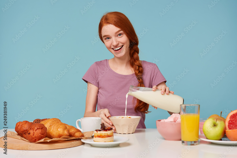 Cheerful kind cheerful red-haired girl sitting at the table is going to have breakfast. Pour milk into a plate with mussels. Smiling affably, looking at the camera. Isolated on a blue background. Cheerful kind cheerful red-haired girl sitting at the table is going to have breakfast. Pour milk into a plate with mussels. Smiling affably, looking at the camera. Isolated on a blue background.