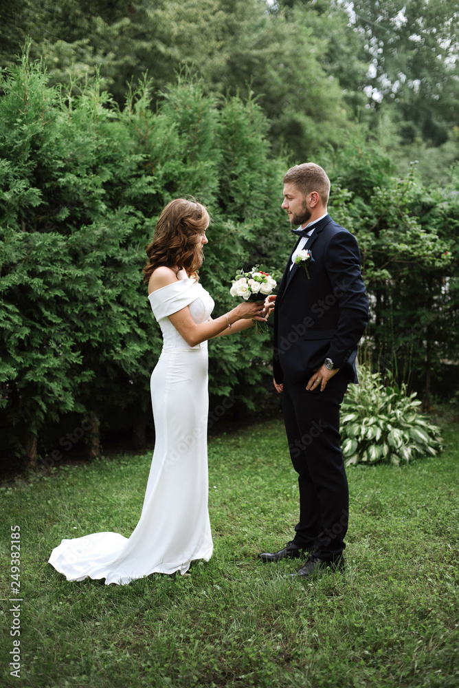 Two young beautiful lovers getting ready for the wedding outdoor on green background. Pretty bride putting on the buttonhole bouquet. wedding concept Two young beautiful lovers getting ready for the wedding outdoor on green background. Pretty bride putting on the buttonhole bouquet. wedding concept