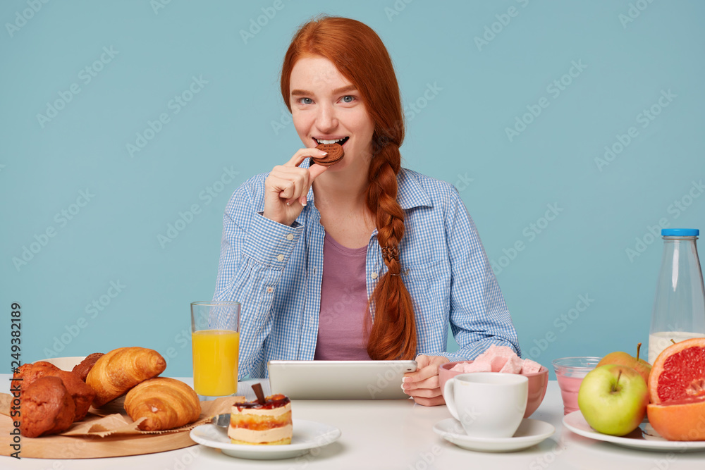 Cute attractive red-haired girl with a smile eating cookies sitting at a table, in her hands holding a tablet on which reads the news, looks into the camera and smiles, against a blue background. Cute attractive red-haired girl with a smile eating cookies sitting at a table, in her hands holding a tablet on which reads the news, looks into the camera and smiles, against a blue background.
