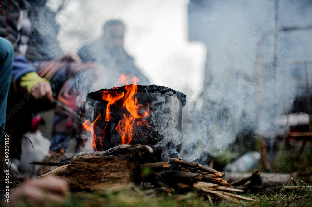 Black pan standing on the campfire on the background of the group of friends Black pan standing on the campfire on the background of the group of friends
