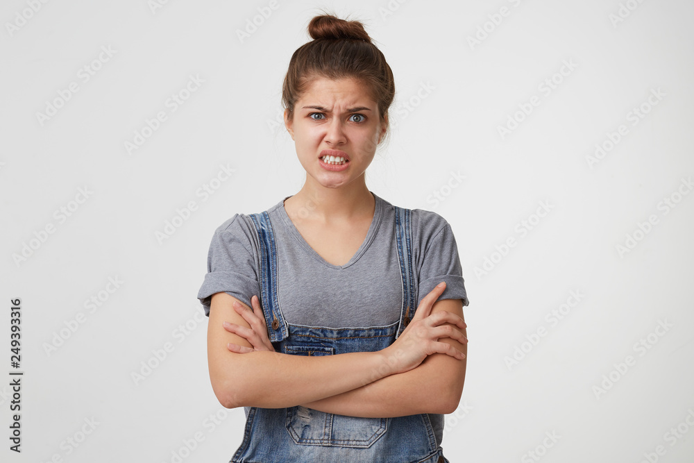 A portrait of a girl who is very angry at her cat or drunk husband. Woman in denim overall shows anger by clasping his teeth and folding her arms, isolated over white background.