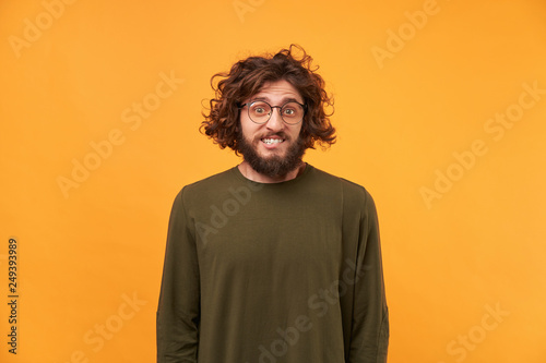 A man with a beard in glasses and curly dark hair looking at the camera, bite lip, confused bewildered as if made mistake, feel guilty isolated on a yellow background dressed in ordinary clothes.
