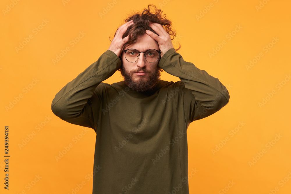 A man in glasses with a beard clutched at his head looks upset sad, got in desperate situations. Doomed exhausted dressed in ordinary clothes, isolated on a yellow background. A man in glasses with a beard clutched at his head looks upset sad, got in desperate situations. Doomed exhausted dressed in ordinary clothes, isolated on a yellow background.