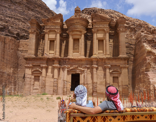 Couple of Male and female tourists in traditional arabic headdress keffiyeh sit on a bench in front of Monastery Ad Deir. in Petra. View from behind photo