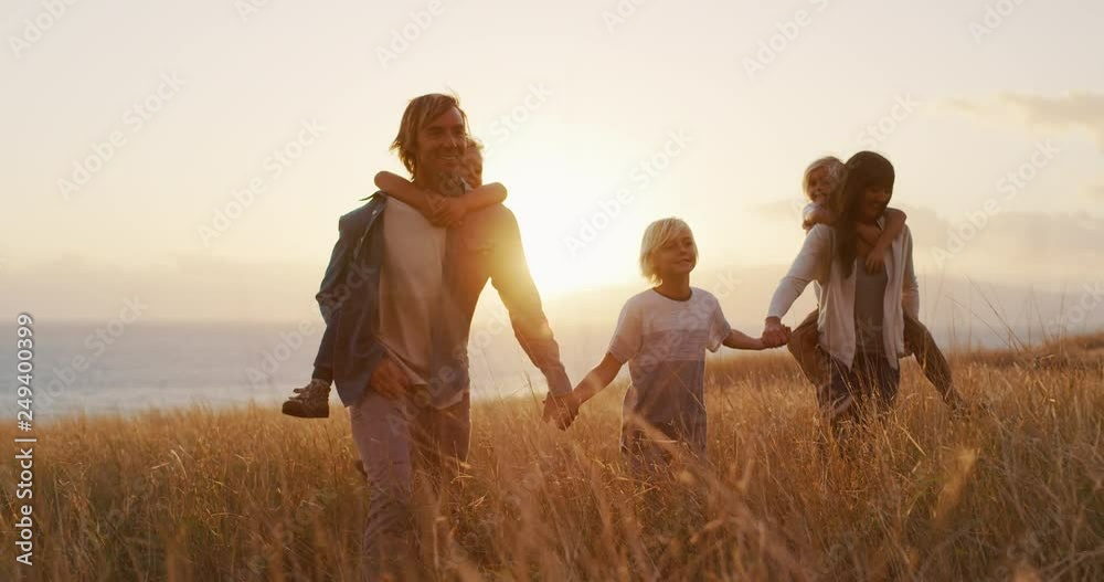 Happy smiling family holding hands walking through golden field at sunset by the ocean, piggy back ride Happy smiling family holding hands walking through golden field at sunset by the ocean, piggy back ride