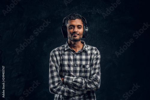 Handsome Indian guy is happy listening to music in wireless headphones standing with his arms crossed in studio against the background of the dark wall