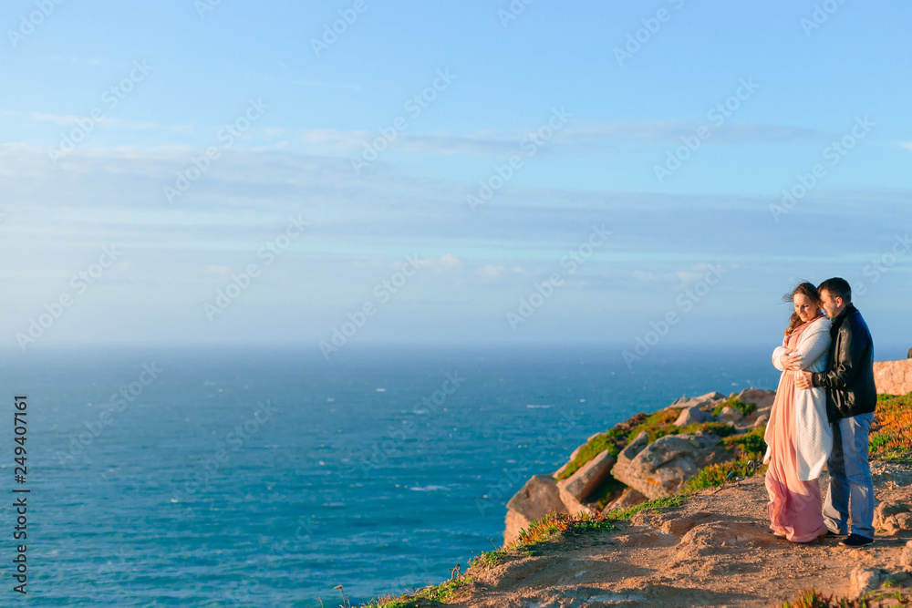 A man hugs a woman from the back side on the rocky shore of the