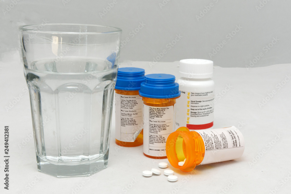 Medicine bottles with caps and one bottle on its side with pills spilling out and a glass of water.