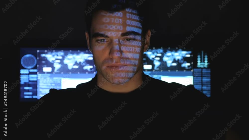 Hacker programmer working on computer in a data center while blue code characters reflect on his face in a dark office room. Hacker programmer working on computer in a data center while blue code characters reflect on his face in a dark office room.