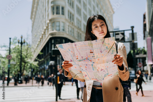 elegant female tourist travel alone in spring holidays to san francisco holding paper map searching direction of famous attraction. white flatiron building in background under sunlight in summer.