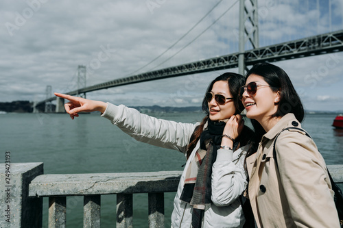 travel woman pointing with finger outdoors and showing something to friend. young backpacker asian sisters tourism in san francisco oakland bay bridge on spring holidays. girls happy seeing nature
