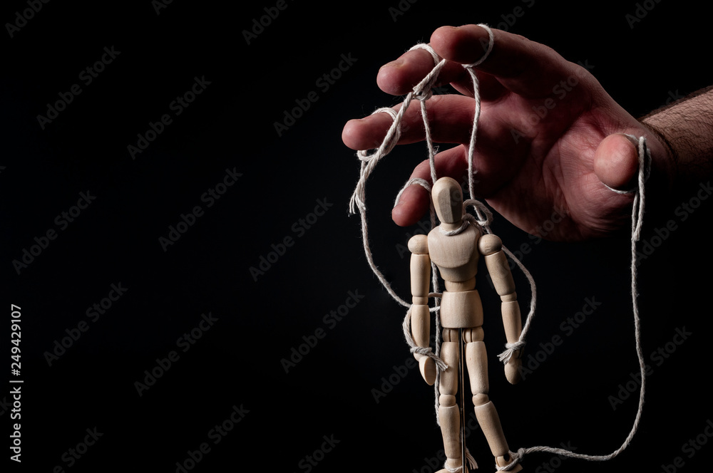 Employer manipulating the employee, emotional manipulation and obey the master concept with ominous hand pulling the strings on a marionette with moody contrast on black background with copy space
