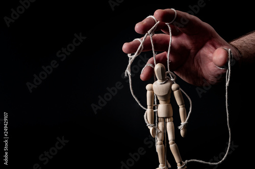 Employer manipulating the employee, emotional manipulation and obey the master concept with ominous hand pulling the strings on a marionette with moody contrast on black background with copy space