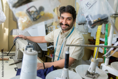 Happy looking tailor working on sewing machine in his workshop with tailoring accessories hanging around.	 photo