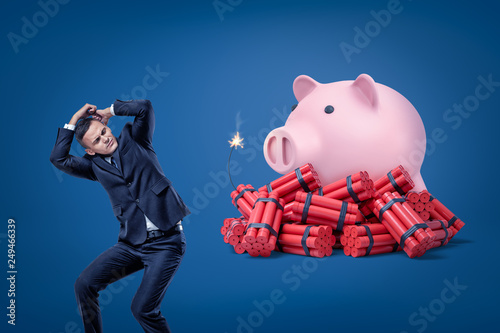 Businessman protecting himself with hands and pink piggy bank over red dynamite sticks with lit fuse on blue background photo