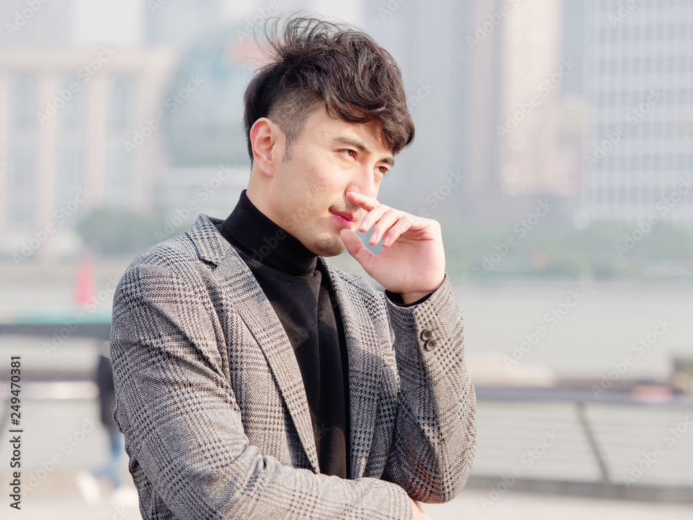 Naklejka premium Portrait of a handsome Chinese young man in casual suit thinking with hand on chin with Shanghai bund background, winter fashion, cool young man lifestyle.