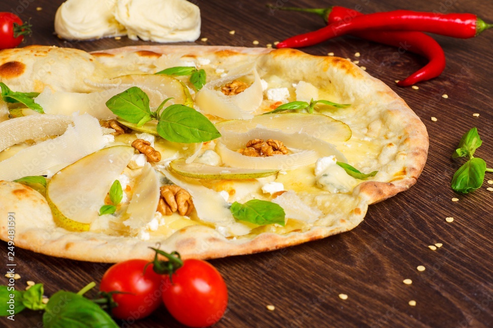 Vegetarian Italian pizza with nuts, yellow pear, parmesan cheese and fresh green basil leaves on a brown table decorated by mozzarella, red hot chili pepper, cherry tomatoes