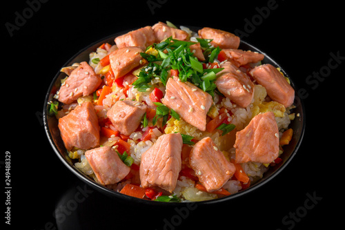 A plate of delicious rice with grilled salmon pieces and vegetables: carrots, sweet peppers, greens, spices and sauce on a black background