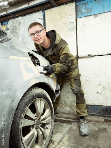 cute guy in black glasses with gloves displays the shape of the hood for the subsequent painting of the car. guy - a house painter working on a car in the garage 