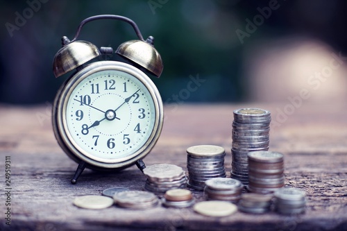 Vintage alarm clock and coin stacks on wooden table with blur green garden background, bright morning color tone, finance and business concept