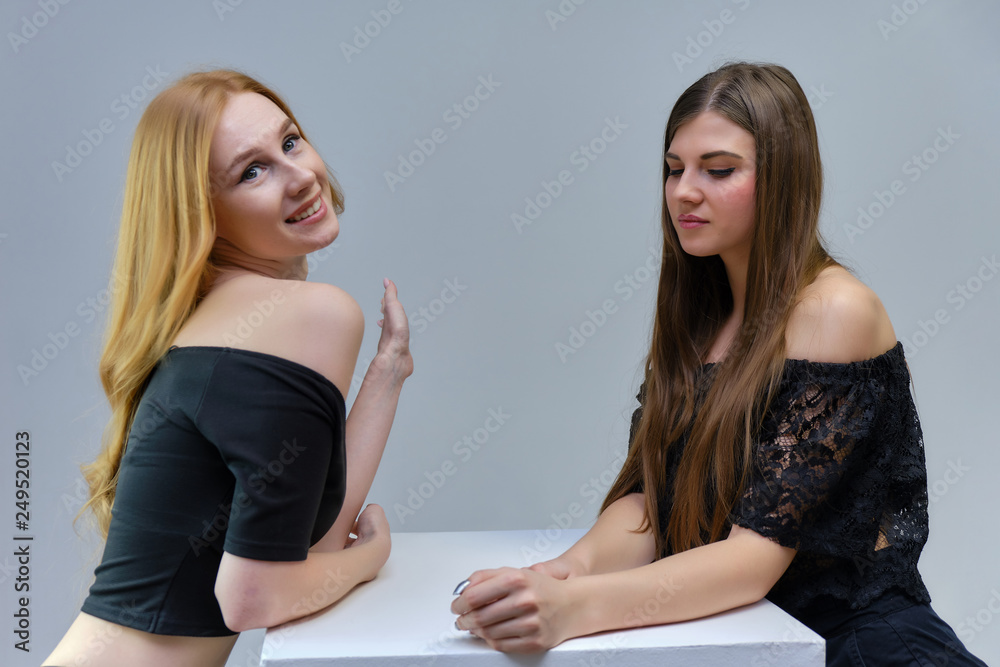 Concept portrait of two cute beautiful girls talking on a gray background in the studio standing at the table Concept portrait of two cute beautiful girls talking on a gray background in the studio standing at the table